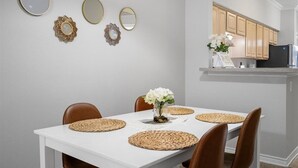 Un comedor con una mesa blanca, sillas marrones y individuales tejidas. Se observan espejos decorativos y obras de arte en la pared sobre la mesa.
