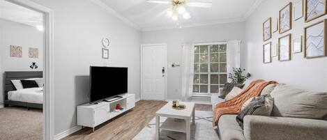 A modern living room with a flat-screen TV, a ceiling fan, and a large window with blinds.