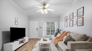 Una sala de estar con una televisión de pantalla plana, un ventilador de techo, un sofá blanco, una mesa de centro y obras de arte enmarcadas en la pared.
