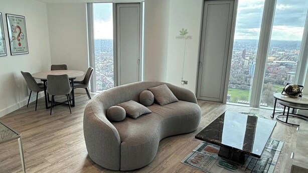 A modern living room with a curved grey sofa, a dining table with chairs, and large windows offering a city view. - 3Bed Apt Sleeps 6 - City Central with Wifi (Manchester)
