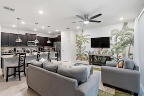 A modern kitchen with dark cabinets, stainless steel appliances, pendant lighting, and a dining area with a gray sofa and chairs.