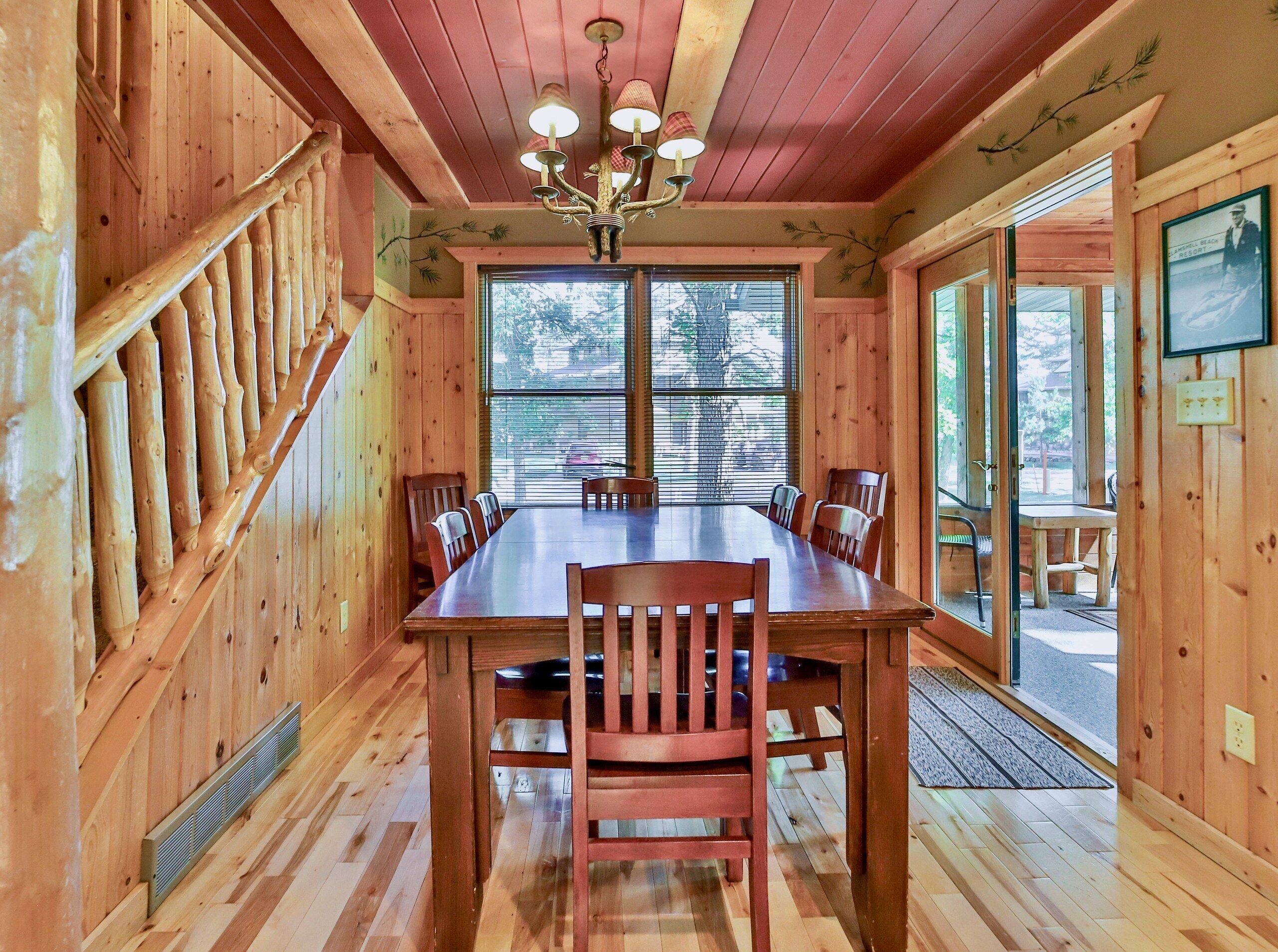 Ein Holzinterieur mit einem Esstisch, Stühlen, einer Chandelieber und großen Fenstern.