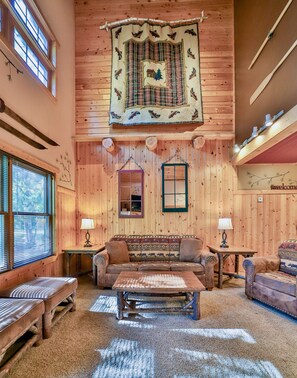 A cozy cabin interior with a large window, a sofa, a wooden table, and a decorative tapestry.