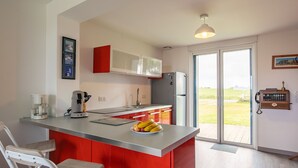 A modern kitchen with a central island, red cabinets, a stainless steel refrigerator, and a large window offering a view of the outdoors. - Le Nid Marin by Interhome (Kerlouan)