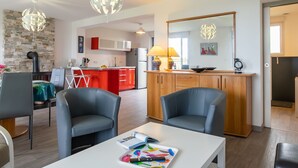 A modern living room with two grey armchairs, a white coffee table with children's books, a kitchen area with red cabinets and a white fridge, and a dining area with a table and chairs. - Le Nid Marin by Interhome (Kerlouan)