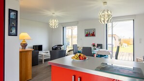 A modern kitchen with a red island, a dining area with a table and chairs, and a living space with a sofa and a TV. - Le Nid Marin by Interhome (Kerlouan)