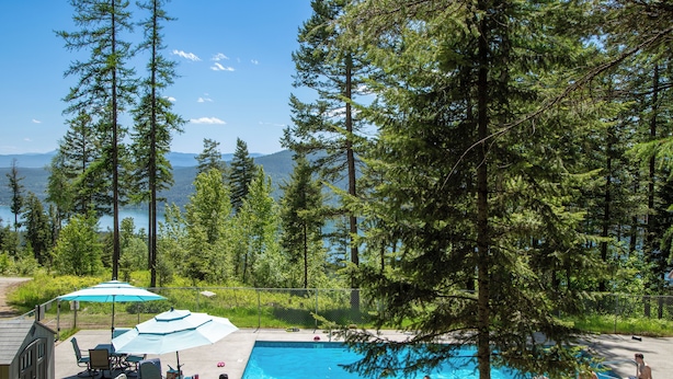 A pool area with a view of a lake and mountains, surrounded by tall trees. - New! Lake Access, Pools/Hot Tub & Ski Area Proximity (Whitefish)