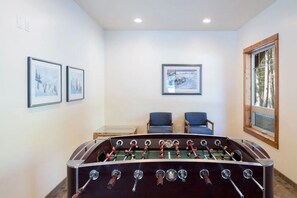 A room with a foosball table, two framed pictures on the wall, two chairs, and a window with a view of trees.