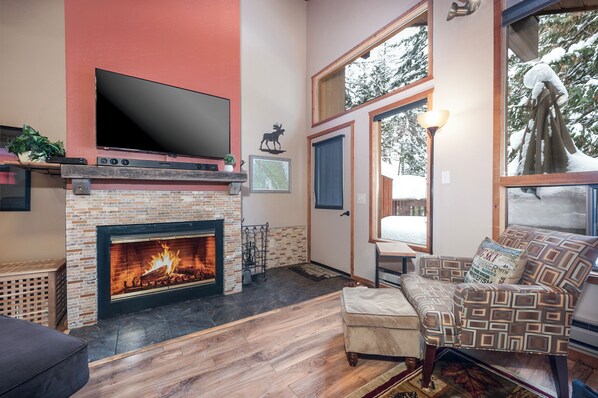 A living room with a fireplace, a television mounted above it, and a large window with a view of snow-covered trees. - New! Lake Access, Pools/Hot Tub & Ski Area Proximity (Whitefish)