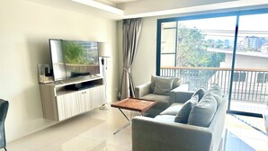 A modern living room with a flat-screen TV, a sofa, a coffee table, and a balcony with a view of buildings and trees. - Le Bus  (Pattaya)
