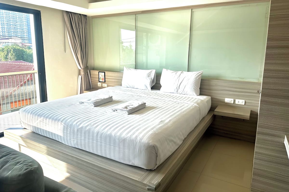A modern hotel room with a large bed, a nightstand, a window with curtains, and a view of buildings outside.