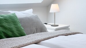 A neatly made bed with a white and beige blanket, a green textured pillow, and a grey ribbed pillow. A white bedside table with a lamp and a black object is visible.