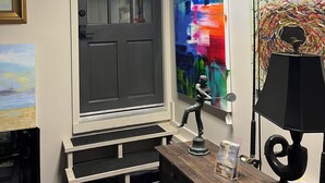 A room with a large window, a door, a desk with a lamp, and various artworks on the walls.