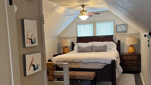 A bedroom with a bed, a ceiling fan, a window with blinds, a bedside table with a lamp, a rug, and a door with artwork.