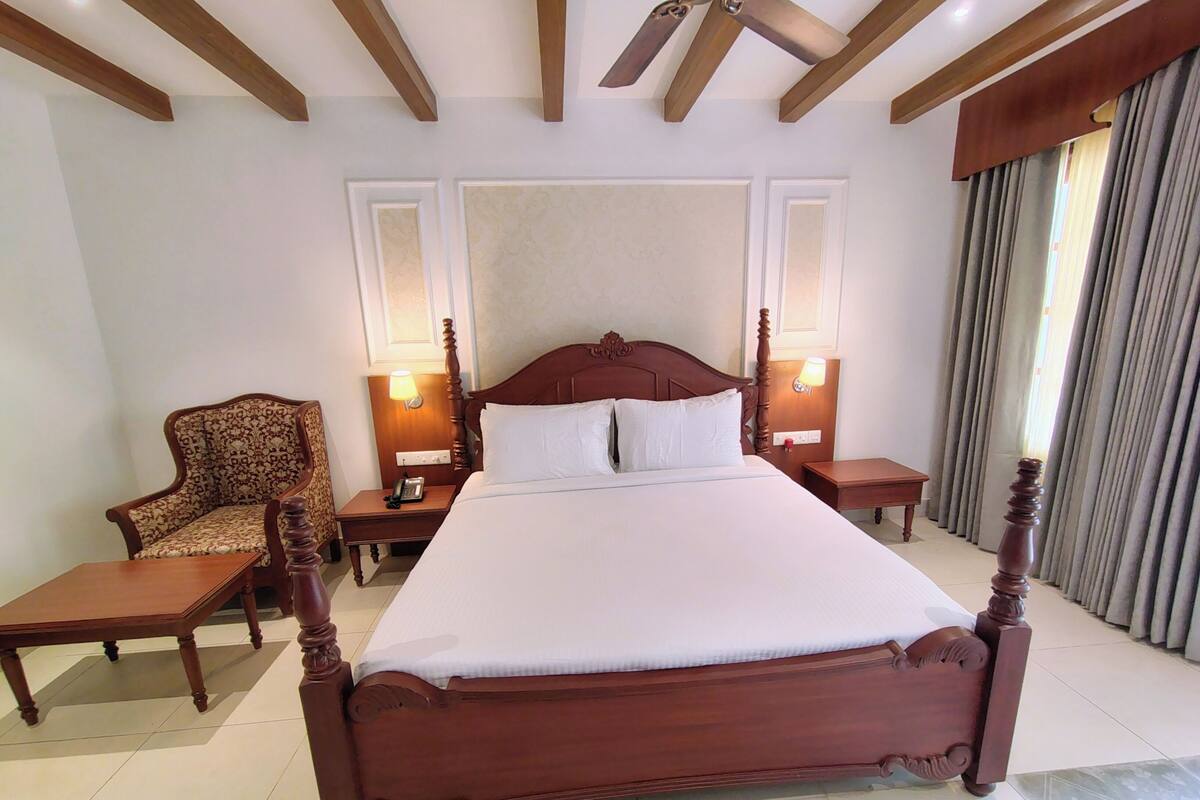 Une chambre d’hôtel avec un grand lit en bois, deux tables de chevet avec des lampes, une chaise en bois et une grande fenêtre avec des rideaux.