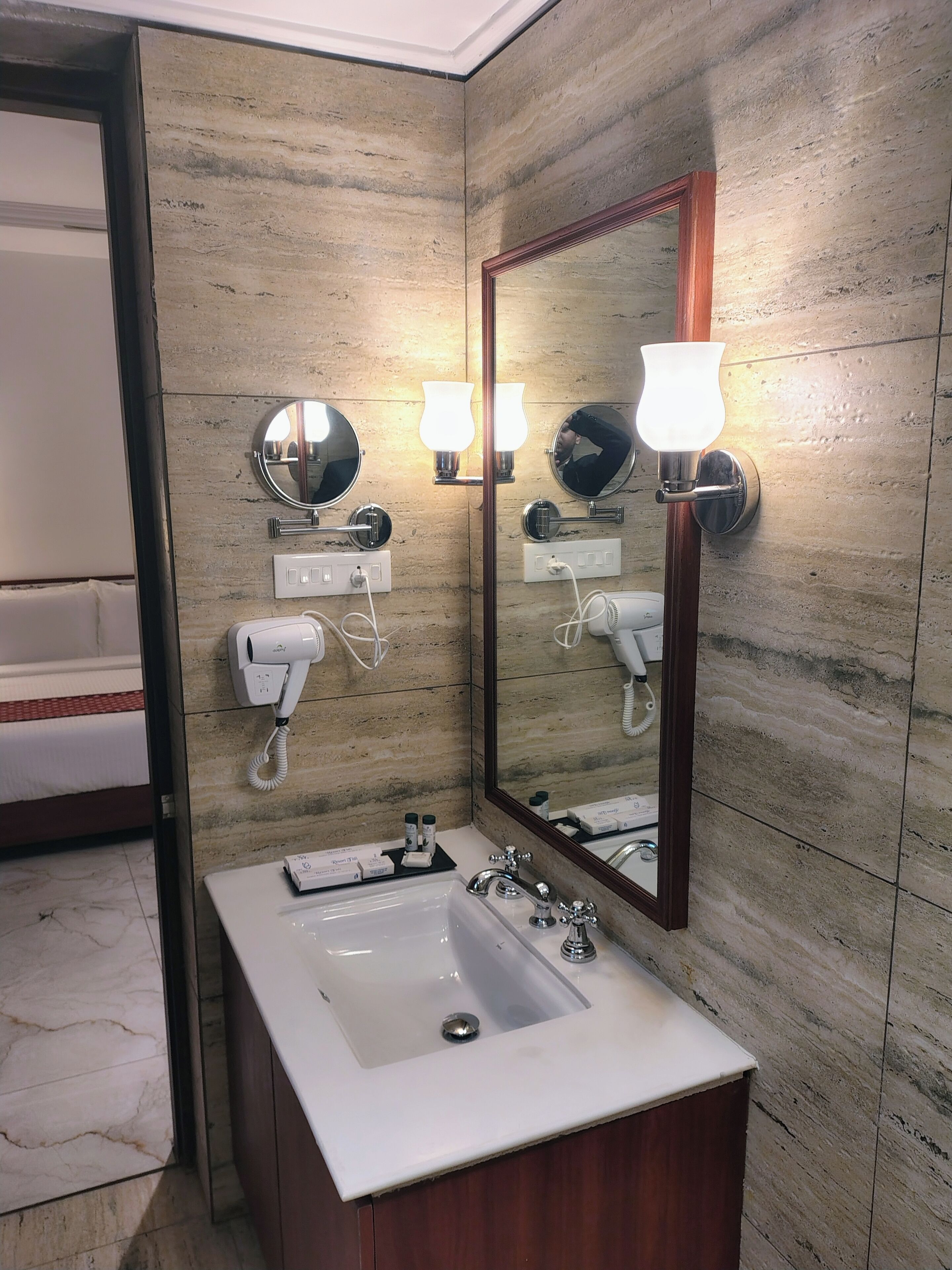 Un baño con lavamanos de mármol, un espejo grande, luces en la pared, un secador de pelo y un perchero para toallas.
