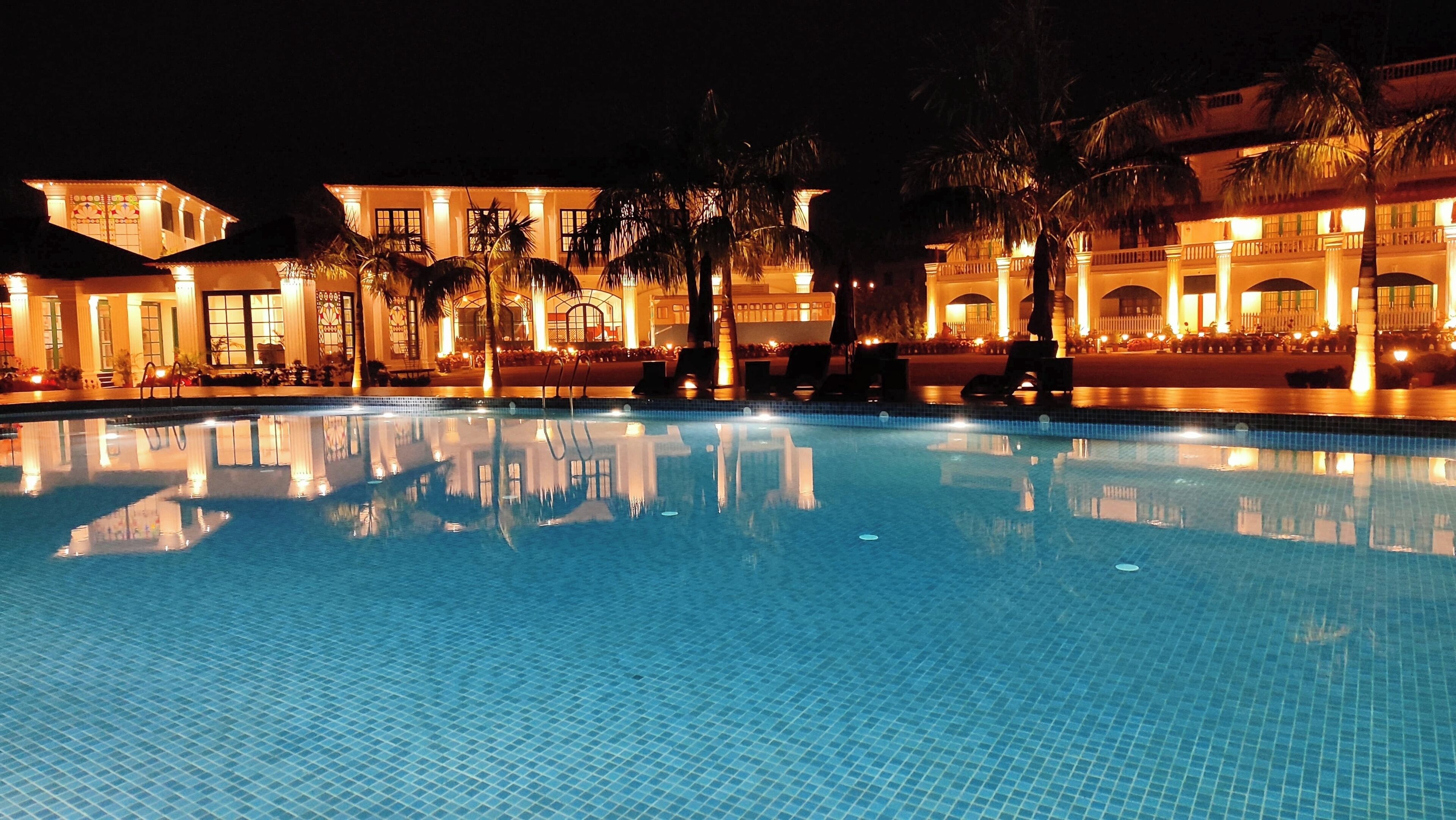 Vista noturna de uma piscina de hotel com água azul cristalina, palmeiras e edifícios iluminados ao fundo.