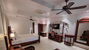 A hotel room with a bed, desk, chairs, a television, a mirror, and ceiling fans. - RESORT TITLI (Bolpur)