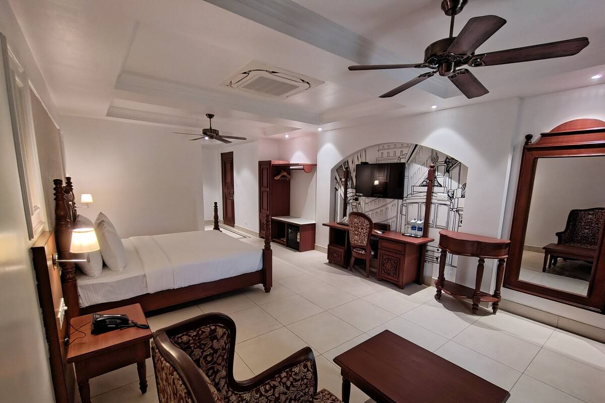 Une chambre d’hôtel équipée d’un lit, d’un bureau, de chaises, d’une télévision, d’un miroir et de ventilateurs de plafond.