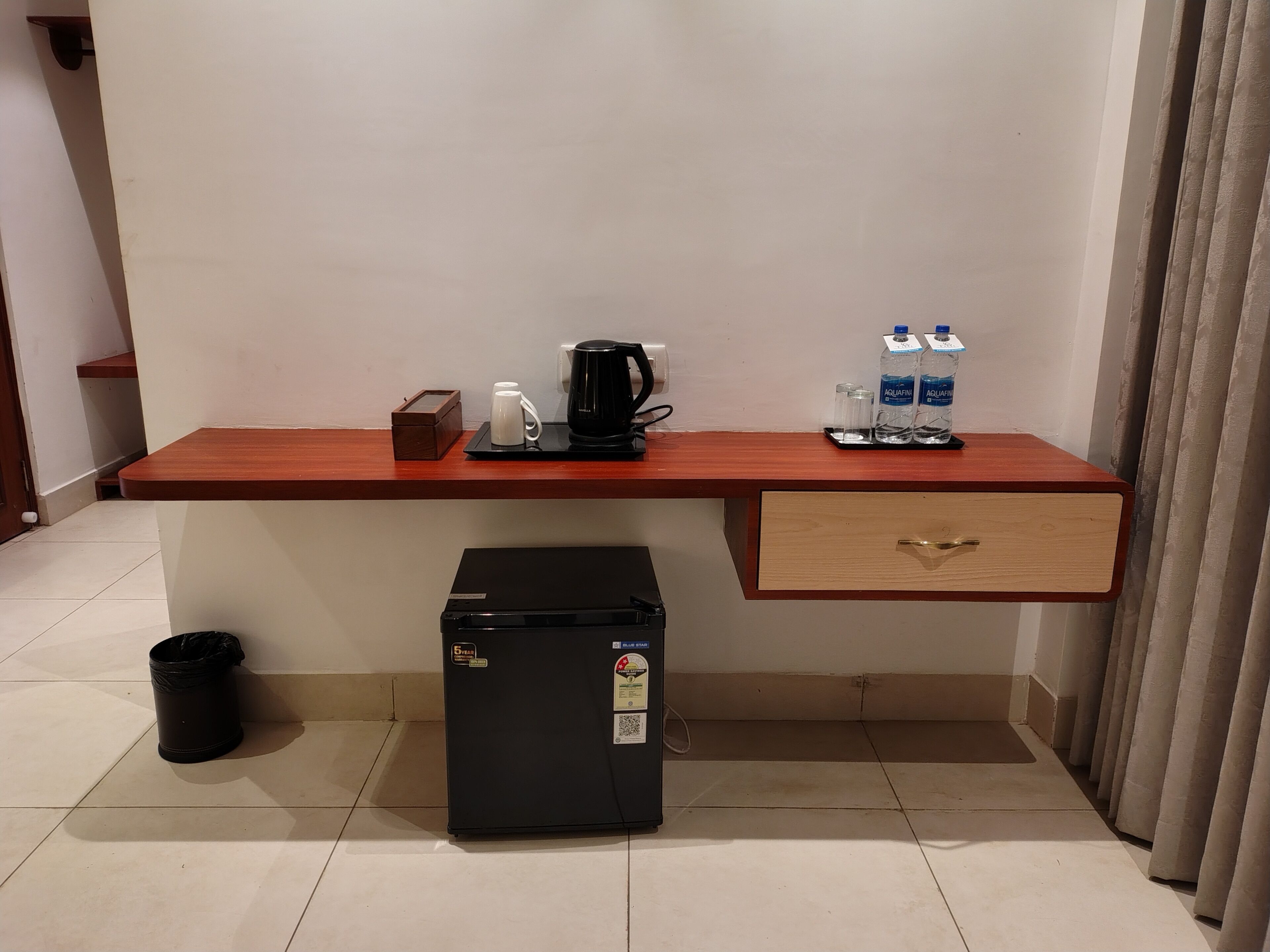 Ein kleines Hotelzimmer mit Schreibtisch aus Holz, kleinem Kühlschrank, Mülleimer, Kaffeemaschine und Wasserspender sowie einer Schublade.