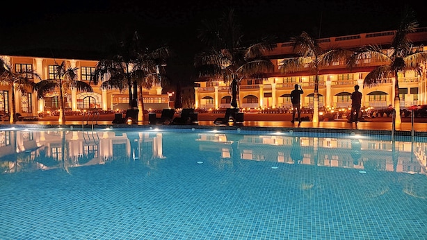 A nighttime view of a hotel pool area with a large pool, palm trees, and a building with multiple balconies. - RESORT TITLI (Bolpur)