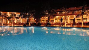 A nighttime view of a hotel pool area with a large pool, palm trees, and a building with multiple balconies. - RESORT TITLI (Bolpur)