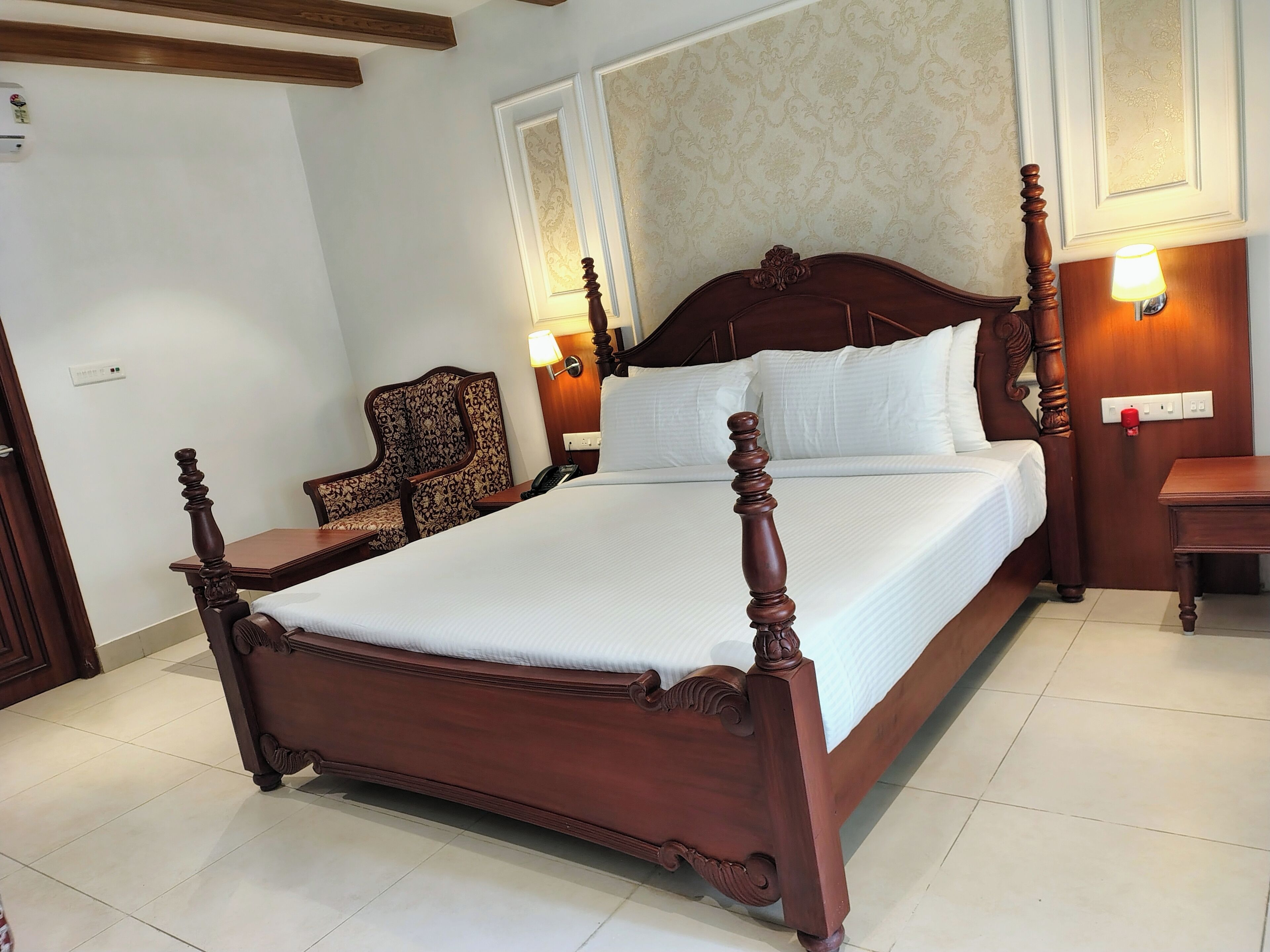 A four-poster bed with a wooden headboard, two bedside lamps, and a chair with a patterned upholstery.