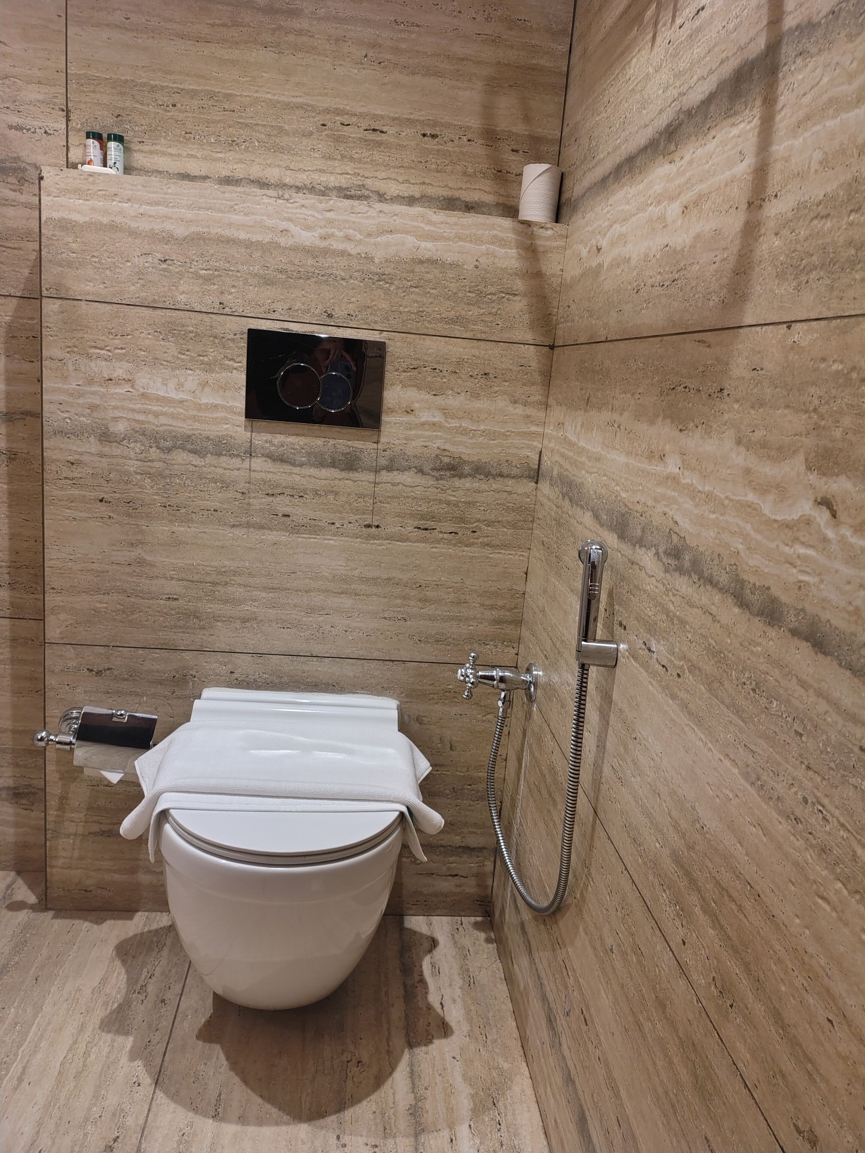 A modern bathroom with a marble wall, a toilet with a towel, a bidet, and a paper towel dispenser.