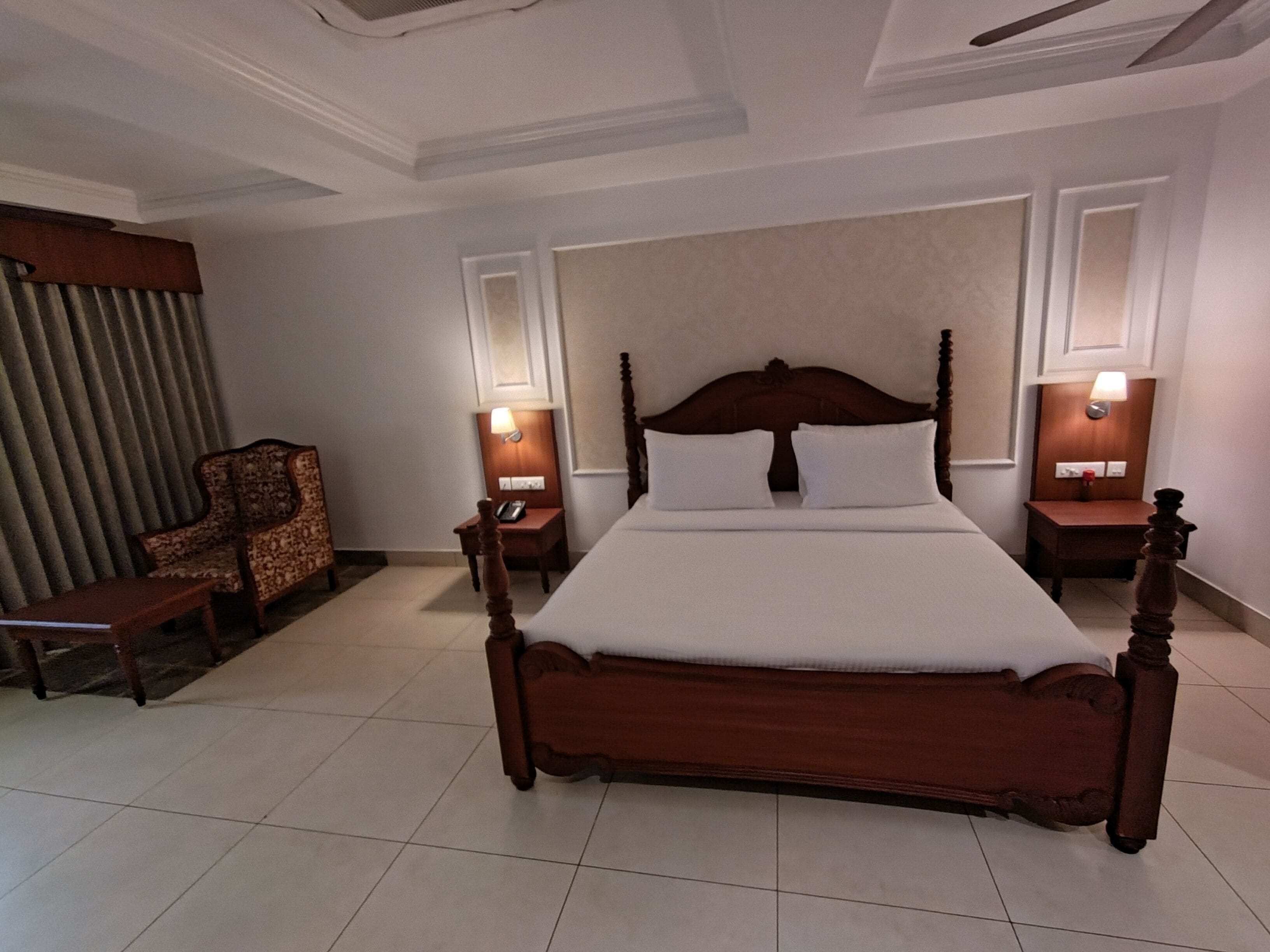 A hotel room with a large wooden bed, two bedside tables with lamps, a wooden chair, and a large window with curtains.
