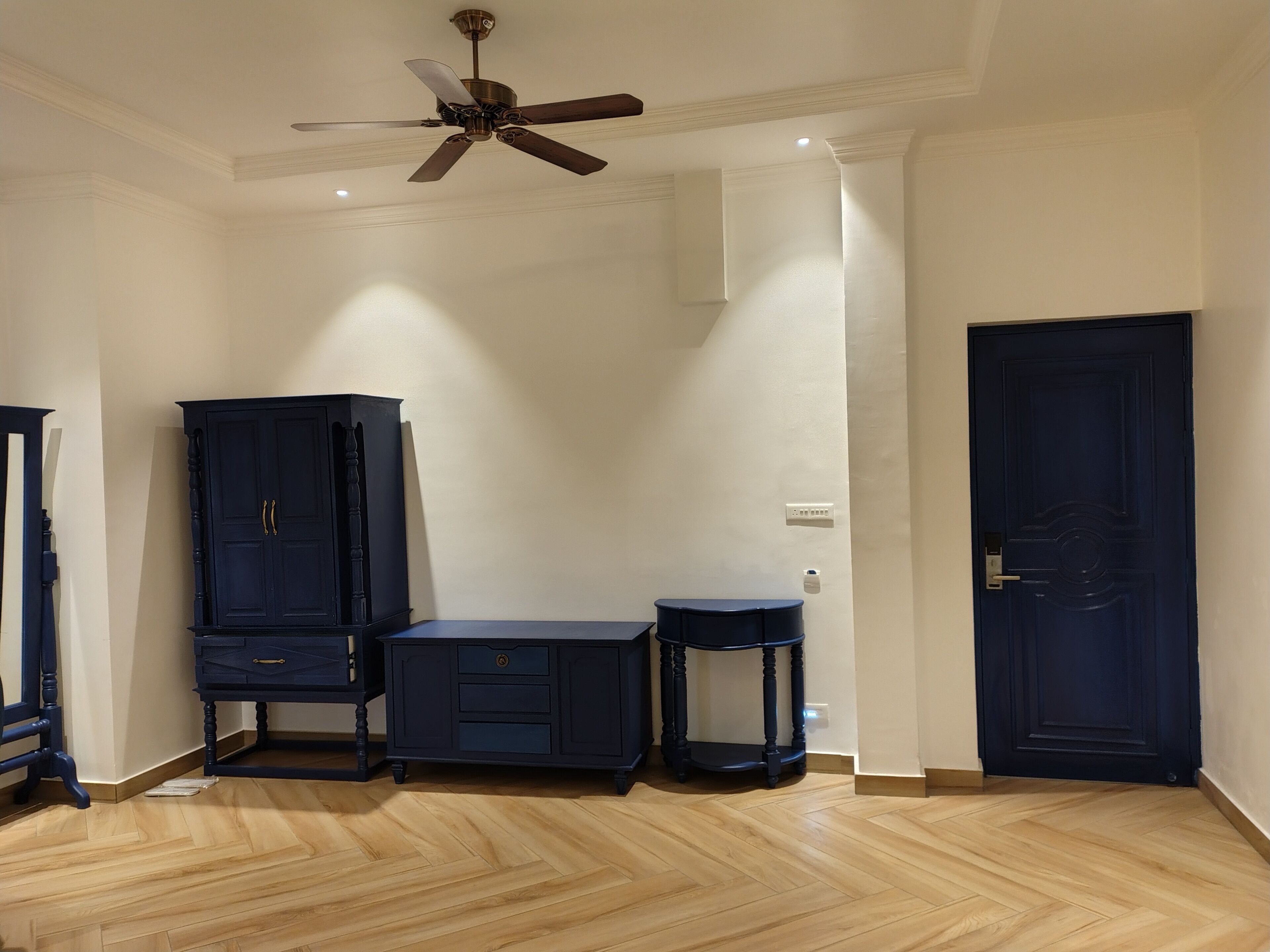 Een kamer met een plafondventilator, een donkerblauwe garderobe, een bureau, een klein rond tafeltje en een donkerblauwe deur.