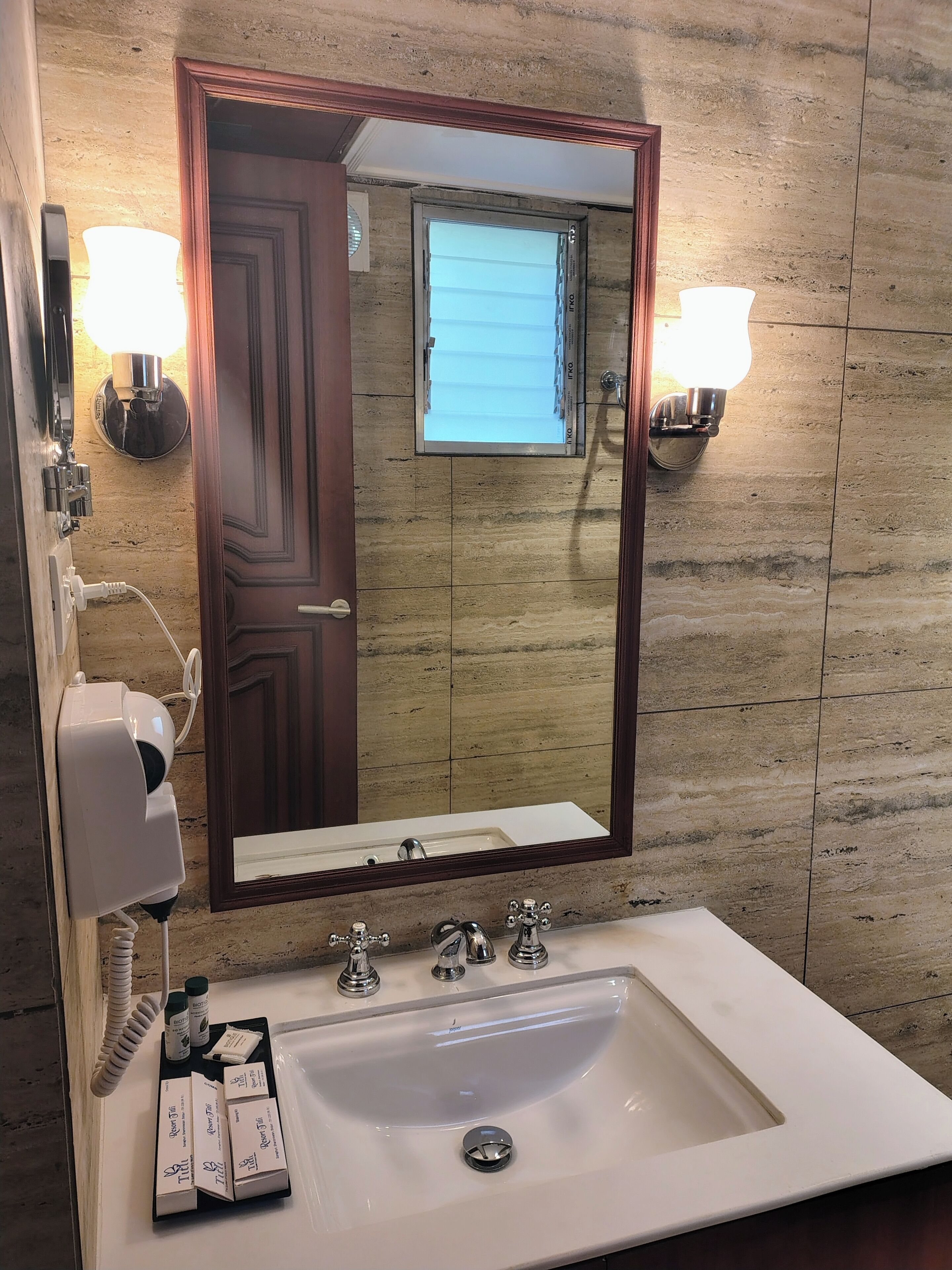 A bathroom with a large mirror, two wall-mounted lights, a hairdryer, and toiletries on the sink.