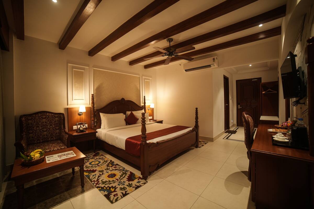 Une chambre d’hôtel avec un lit en bois, un coin salon comprenant une chaise et une petite table, un bureau avec une chaise et un téléviseur fixé au mur.