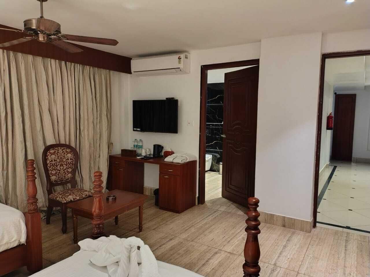 A hotel room with a bed, wooden furniture, a desk, a chair, a television, an air conditioning unit, and a view of the outside.