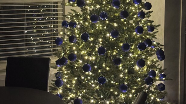 Un sapin de Noël décoré d’ornements et de guirlandes lumineuses bleues, placé dans une pièce où se trouvent une table à manger et des chaises.