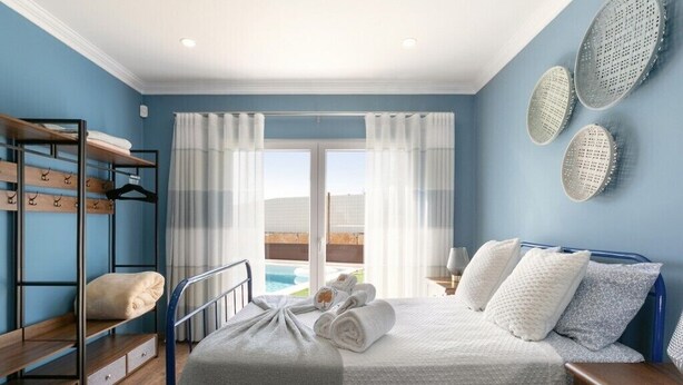 Une chambre avec un lit superposé, un lit recouvert d’une couverture grise, une table de chevet avec une lampe, et une vue sur une piscine par la fenêtre.