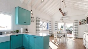 Une cuisine avec des armoires de couleur turquoise, un comptoir blanc et un plancher en bois. On y trouve un coin repas avec une table et des chaises, et une vue sur l’extérieur grâce à de grandes fenêtres.
