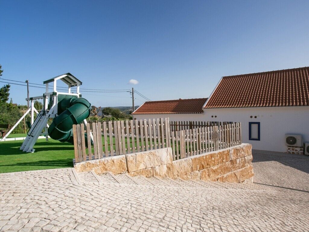 A playground with a slide and a climbing structure, a brick pathway, and a building with a tiled roof.