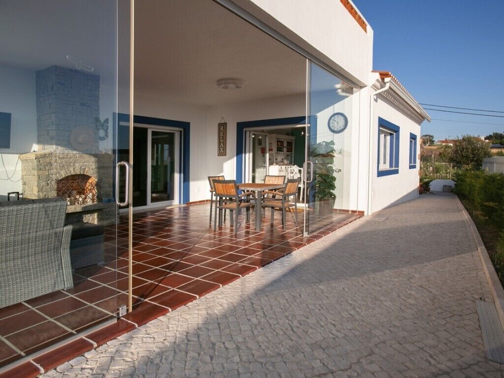 Alpendre coberto com mesa e cadeiras, lareira de pedra e porta de vidro que dá para o interior.