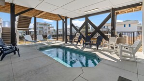 A covered outdoor pool area with lounge chairs and a table, surrounded by a wooden pergola and a view of neighboring houses.