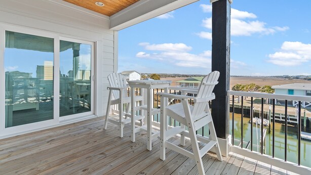 A wooden deck with white chairs and a table, large windows, and a view of a marina.