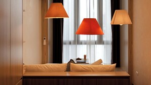 Een moderne kamer met een houten bank, twee oranje hanglampen en uitzicht door een groot raam met dunne gordijnen.