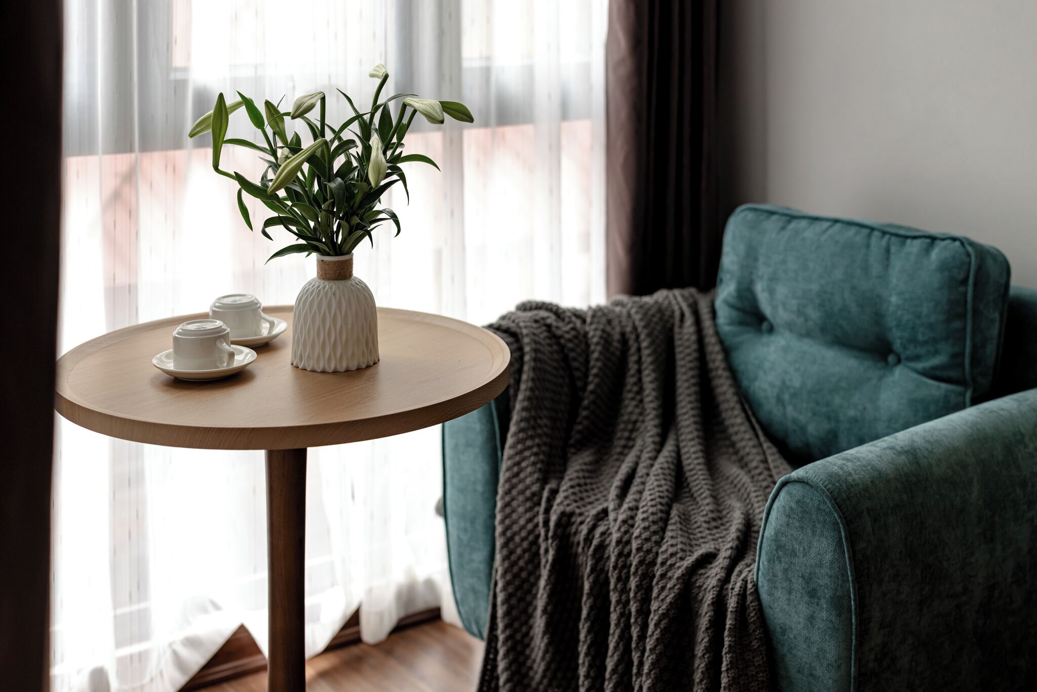 A round wooden table with a white vase and two white cups, a green armchair, and a window with sheer curtains.