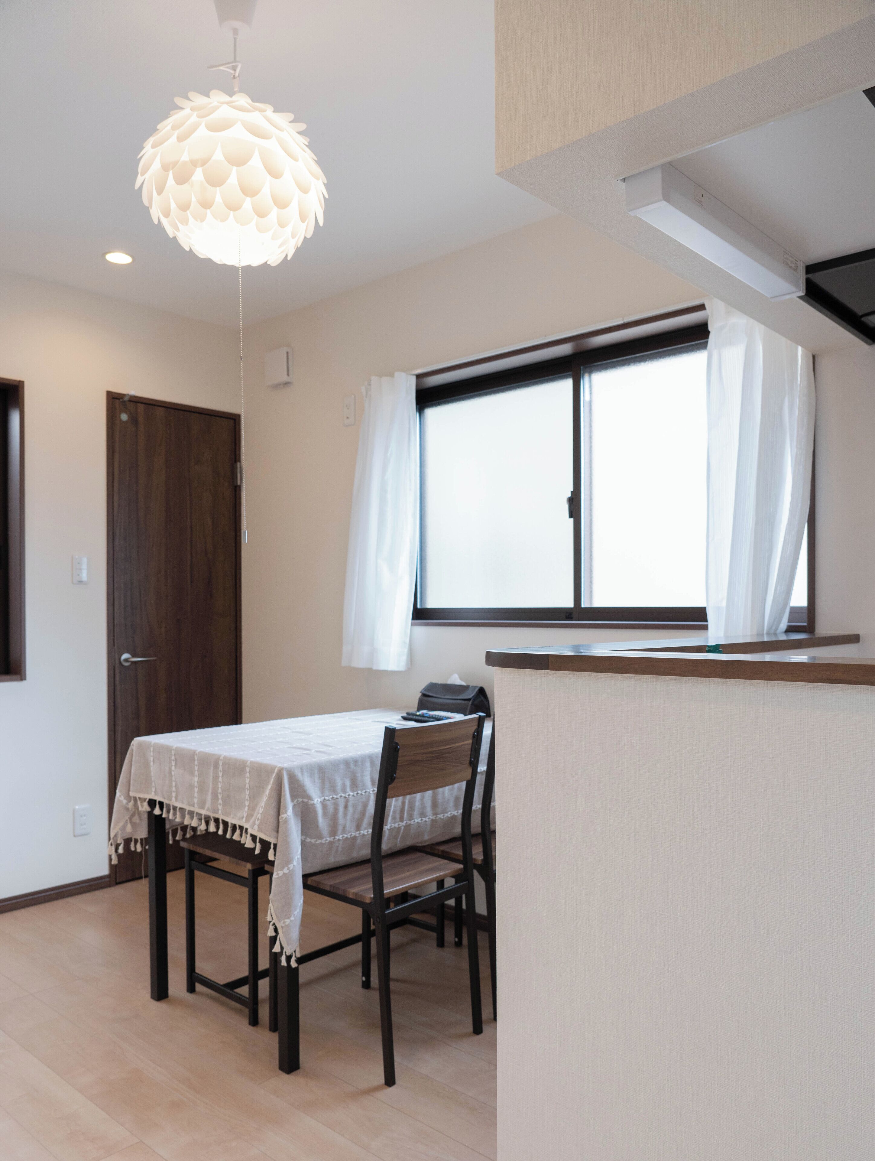 Een kamer met een eettafel, stoelen, een hanglamp, een raam met gordijnen en een deur.