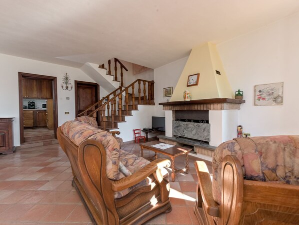 Uma sala de estar ampla com escada de madeira, lareira e área de cozinha visível através de uma porta aberta.