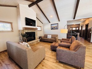 Uma sala de estar ampla com lareira de pedra, uma TV de tela plana montada acima e uma grande janela que oferece iluminação natural.
