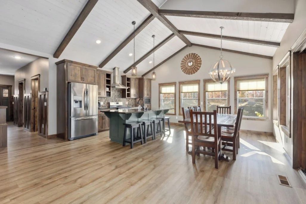 A spacious kitchen with a high ceiling and wooden beams, a central island with bar stools, and large windows providing natural light.