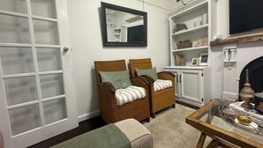 Een woonkamer met rieten stoelen, een glazen salontafel, een open haard en een witte kast met planken.