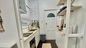 Een smalle keuken met witte kasten, een witte bakstenen achterwand en een witte deur met een ruit. Op de donkerhouten vloer liggen twee beige matten.