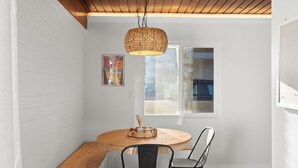 A dining area with a round wooden table, two black chairs, a woven pendant light, and a window with a view of the outside.