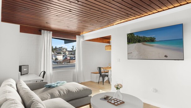 A modern living room with a large window, a sofa, a dining area, and a beach view painting.
